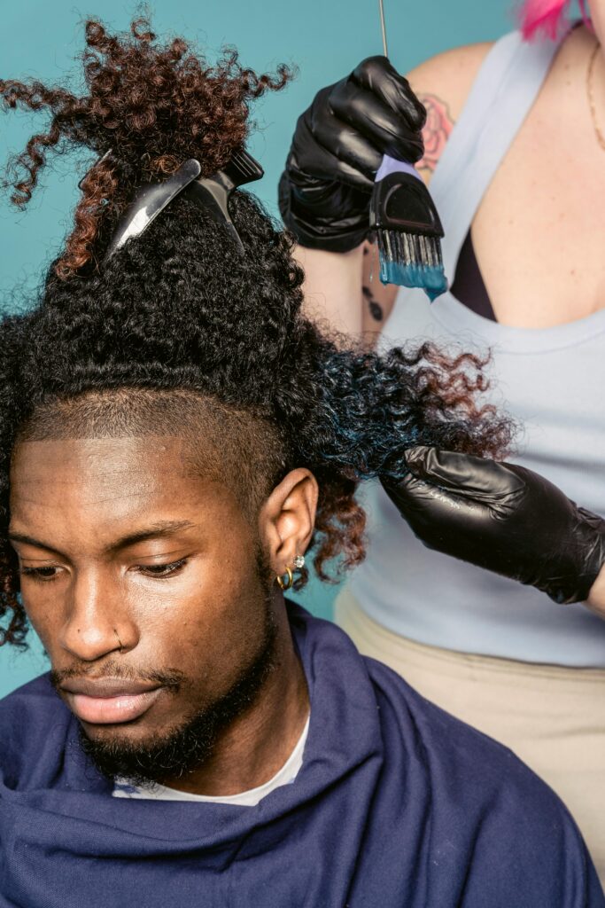pexels-photo-7389077-7389077 A hairstylist wearing gloves applies blue dye to a man's curly hair in a salon.